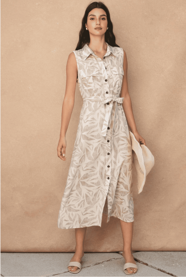 A woman models a sleeveless midi shirt dress with a beige and off-white botanical pattern, featuring a button-down front, collar, and matching waist tie. She is standing against a textured beige wall holding a light beige straw hat.